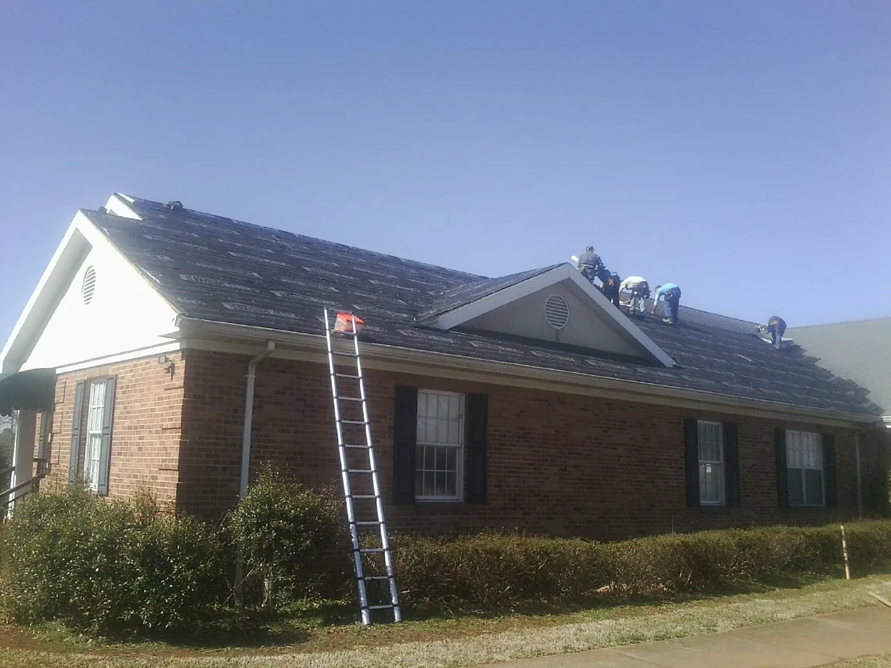 Crew tearing shingles off a brick ranch roof with a ladder set — Metal Roof Repair work by our crew in Reed