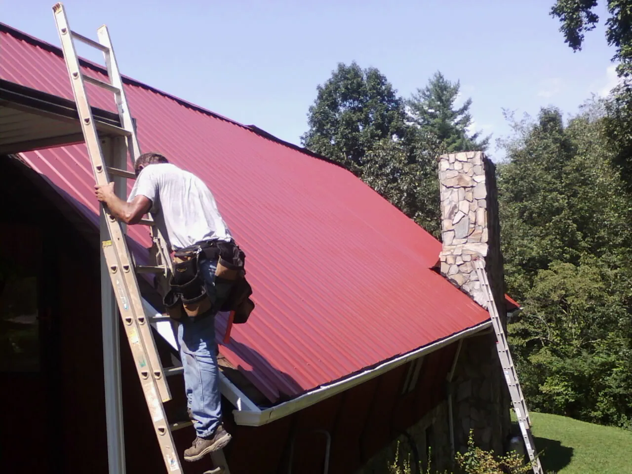 Roofer on a ladder beside a red metal roof and stone chimney — Soffit & Fascia Repair by Roofing Brothers in Reed